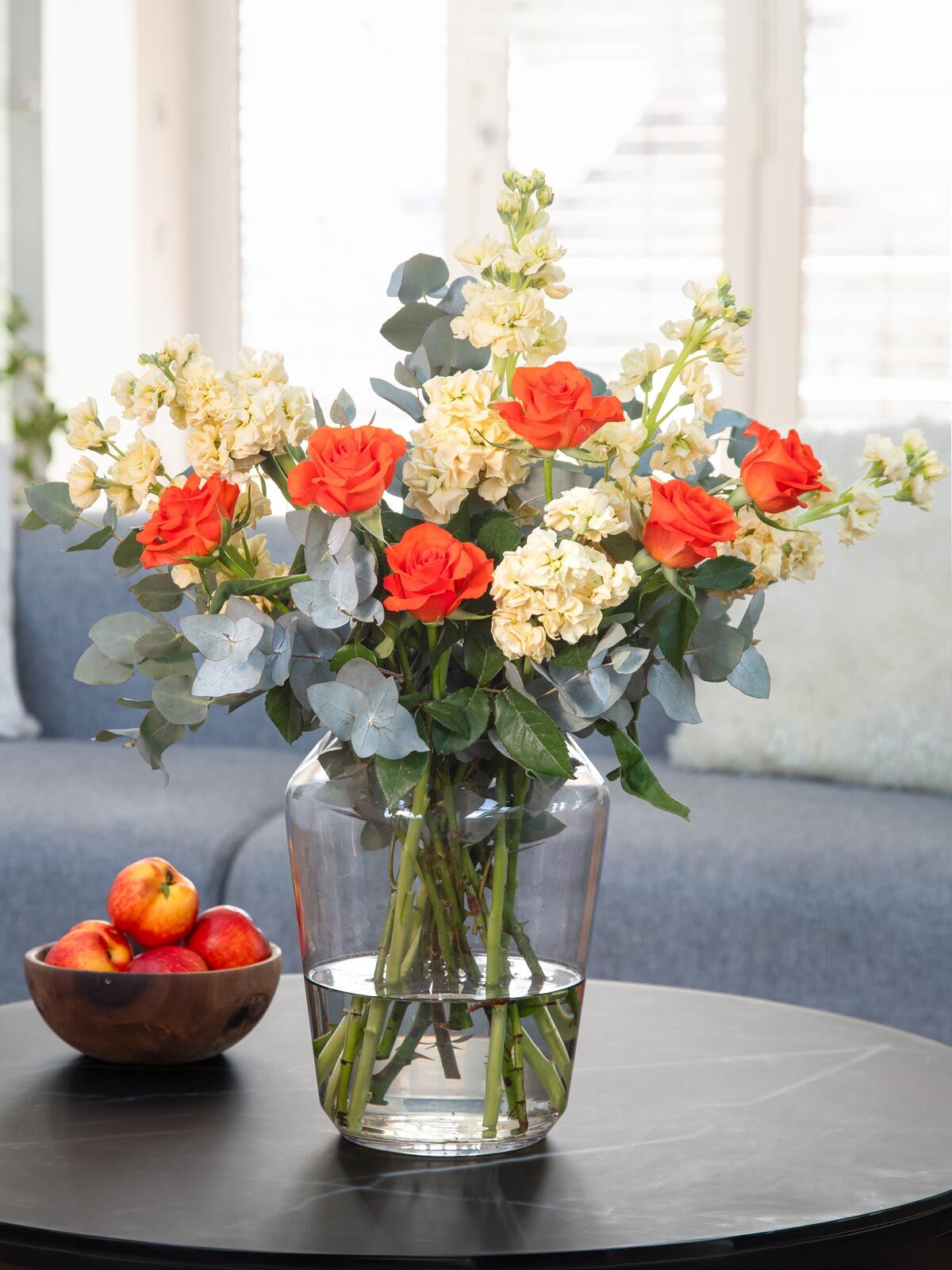 Weiße Levkojen stehen arrangiert mit orangefarbenen Rosen und Eukalyptus arrangiert in einer großen Glasvase auf einem Wohnzimmertisch