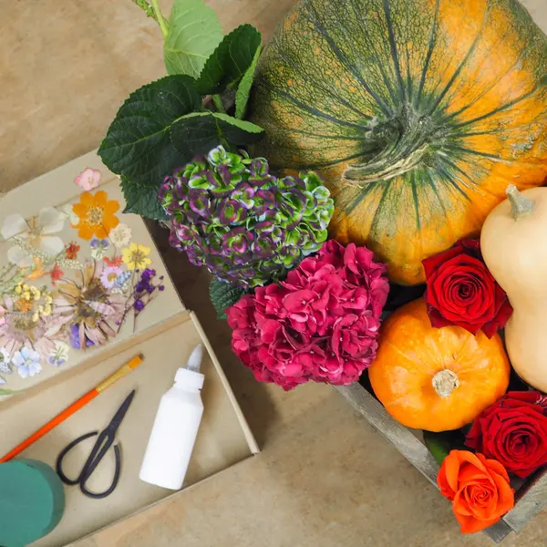 Bastelkleber,Pinsel, Schere, gepresste Blüten in einer Pappbox auf dem Tisch, neben einem Kürbis und Hortensien, sowie Rosen