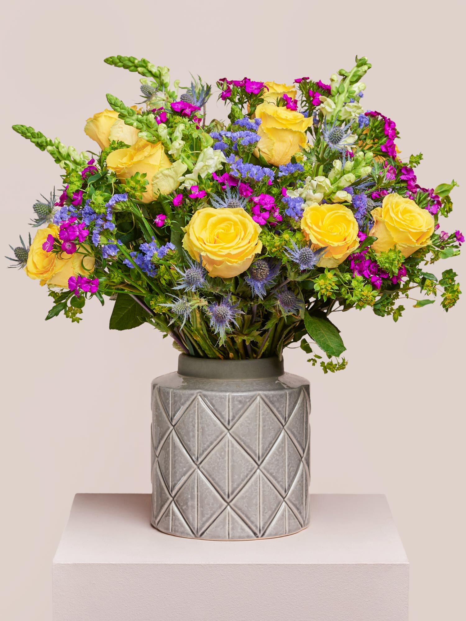 Von allem das Beste Ein großer Blumenstrauß, bestehend aus großen, gelben Rosen, weißen Levkojen, blauen Disteln und Statice, Bubleurum und Bartnelken, arrangiert in einer grauen Keramikvase