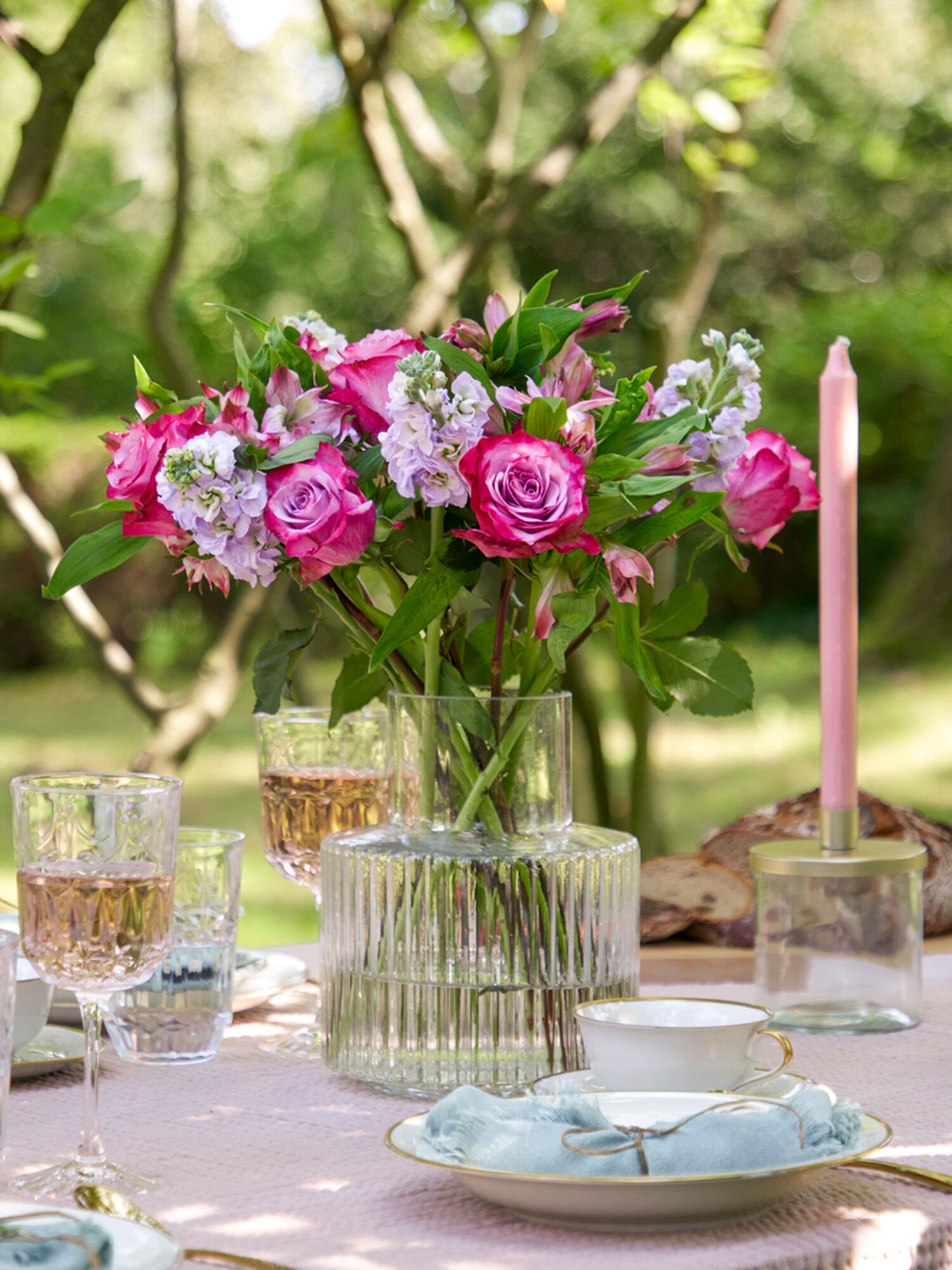 Nahaufnahme eines gedeckter Geburtstagstisch im Garten, mit einer Glasvase, in dem sich ein Blumenstrauß mit pinken Rosen und lilafarbene Levkojen steckt, die Fairtrade sind