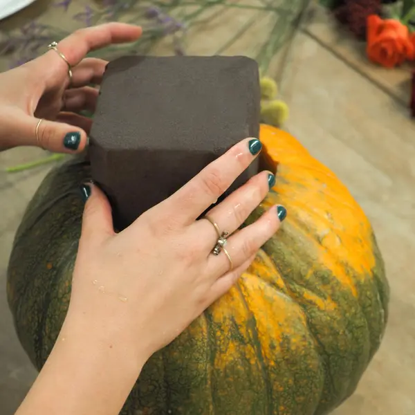 Hände, die einen Würfel Steckschaum in einem ausgehöhlten Kürbis stecken, auf einem Tisch