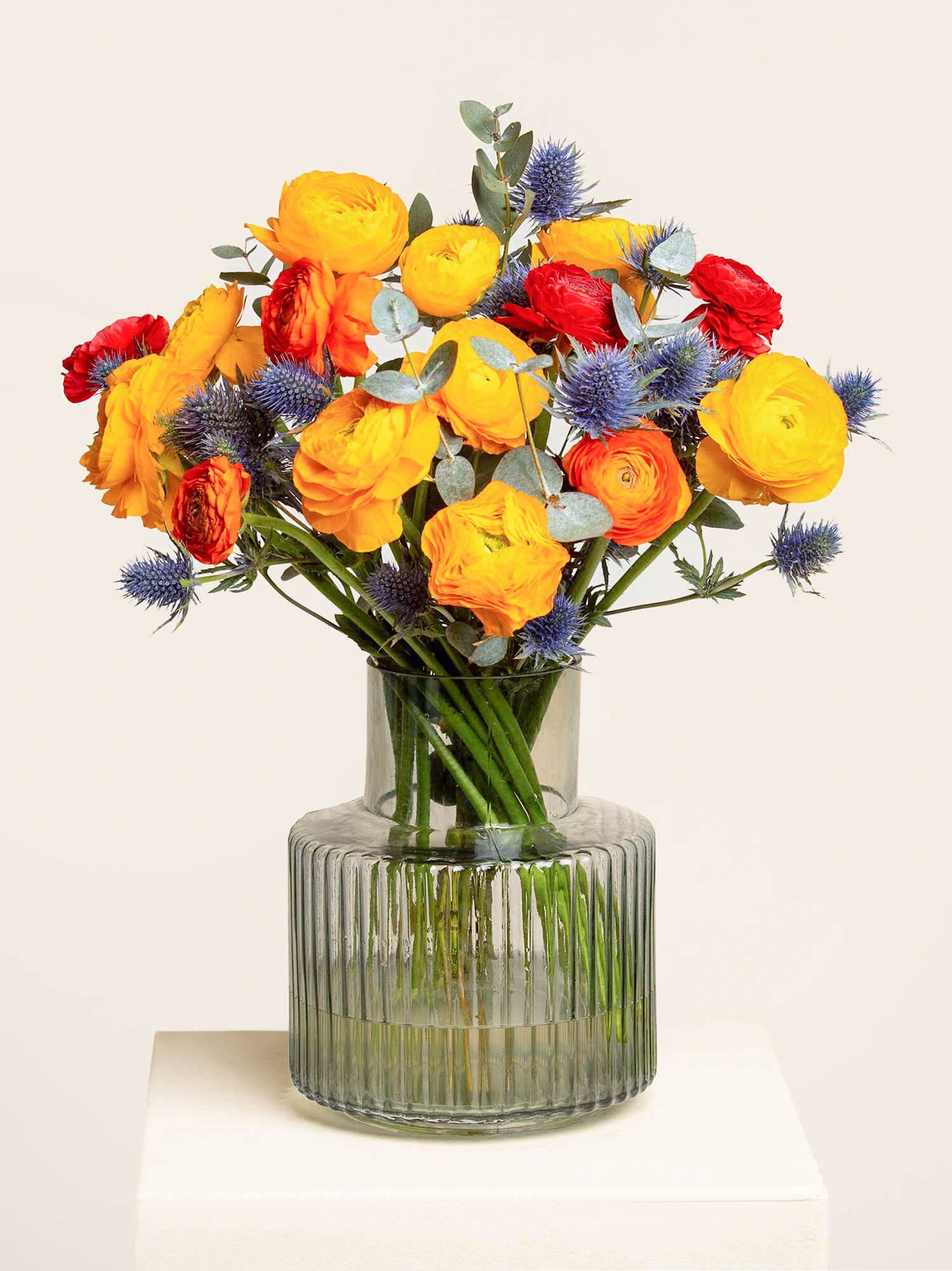 Ein frühlingshaftes Arrangement, bestehend aus gelben und orangefarbenen Ranunkeln, blauen Disteln und Eukalyptus arrangiert in eine Glasvase